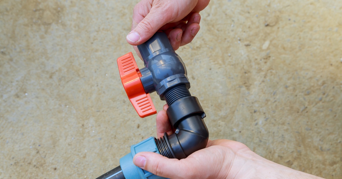 Two hands work to install a red plastic ball valve onto a length of pipe. The pipe is held over a concrete floor.