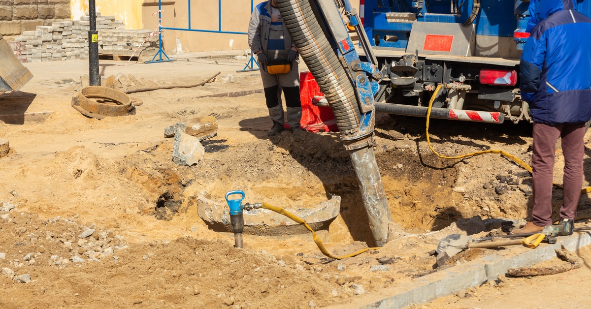 Two workers supervise as a long hose connected to a blue truck excavates a pile of dirt at a construction site.
