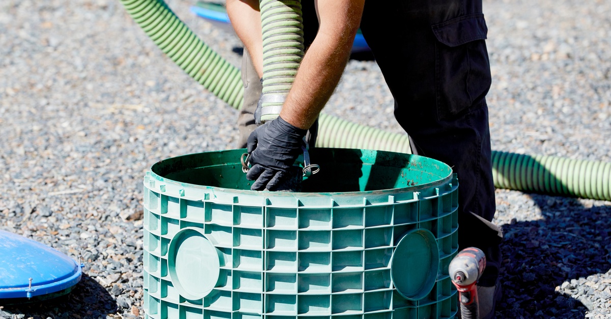 A sanitation worker in work pants is feeding a large hose down a hole in the ground that leads to a septic tank.
