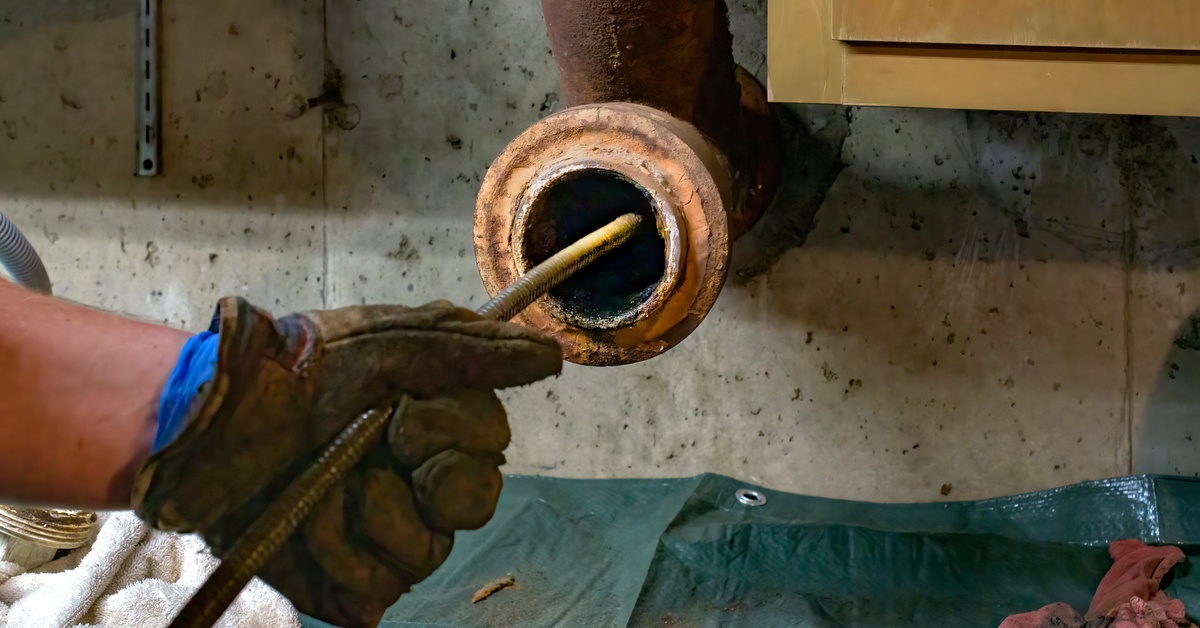 The hands of a plumber as they feed a hose into a rusty drain pipe. The pipe is attached to a dirty concrete wall.