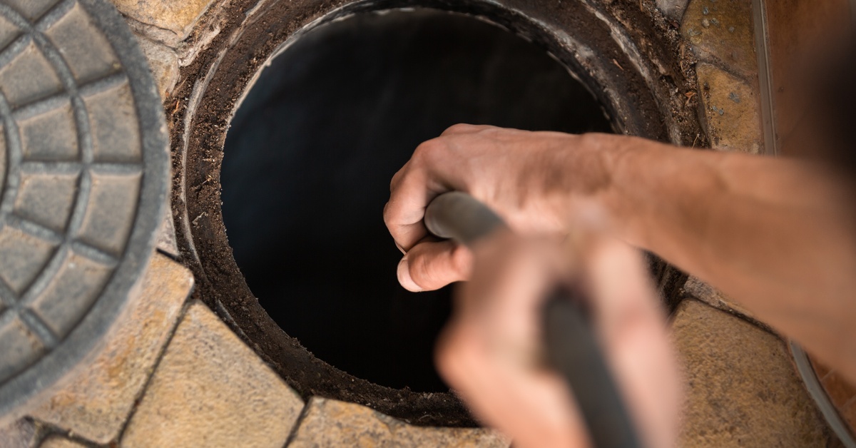 A close up of a plumber's two hands that are feeding a thin hose down a manhole that has its cover off to the side.