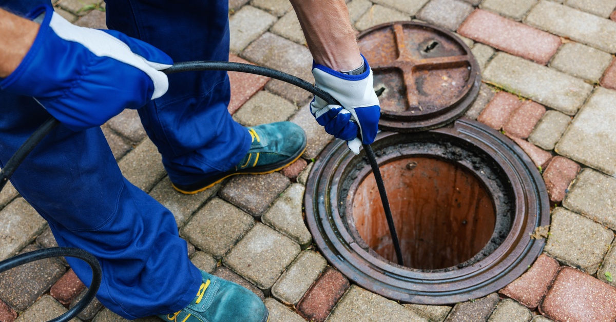 A sewer cleaning professional feeds a thin black hose down a grime-covered hole in a brick layered road.