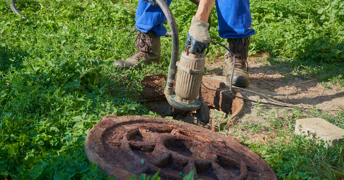 A worker placing a vacuum drainage pump into a cesspool in the ground. The pool is surrounded by grass.