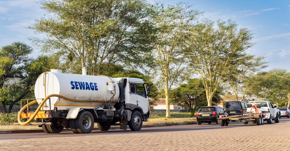 A white sewage truck parked on a brick road. There are several other cars parked in front of the truck.