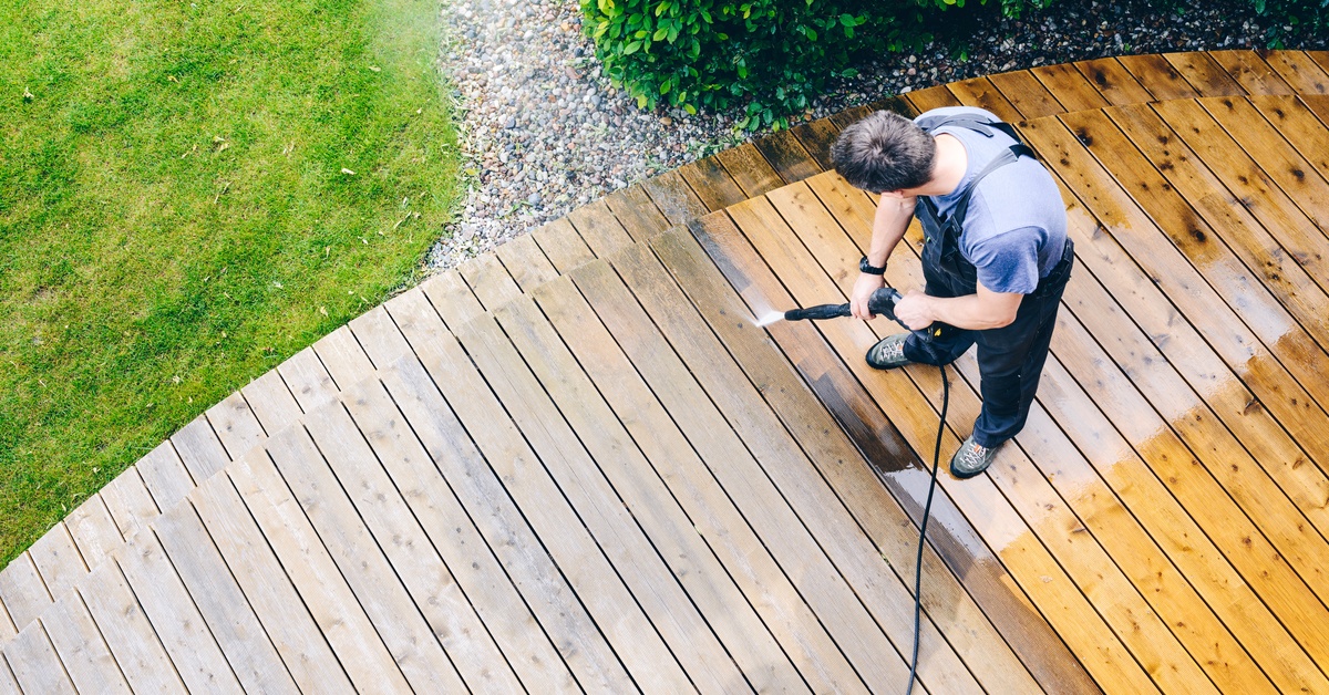 A man with a pressure washer uses it to clean the wooden deck he's standing on. Half of the wood is still dark with dirt.