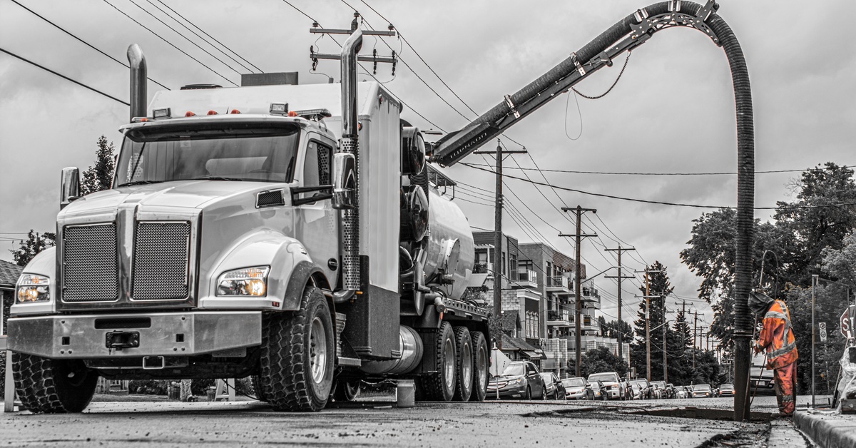A white hydrovac truck has its hose deployed over a hole in the road. The hose is next to a worker in hi-vis clothes.