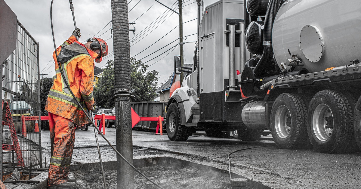 A worker in orange high-visibility clothing guides a large hose into the ground. The hose is connected to a silver truck.