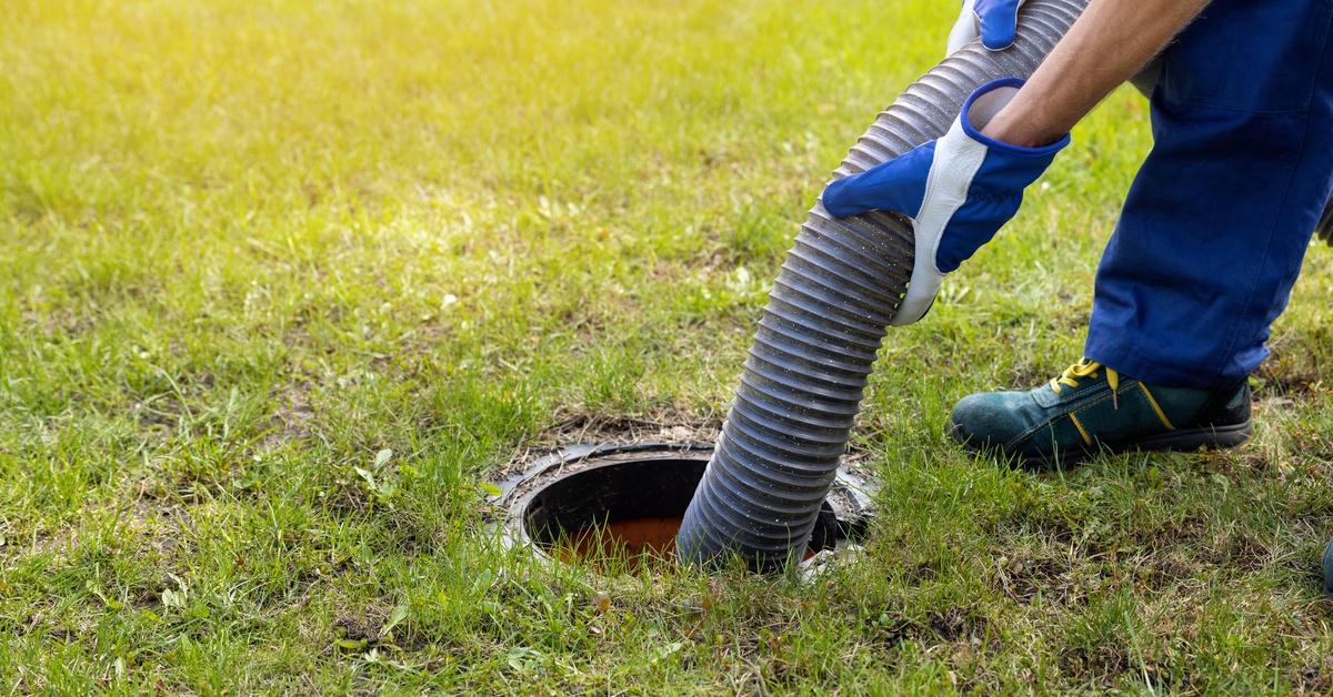 A man wearing gloves is snaking a large hose down into a septic tank that is built into the ground around it.