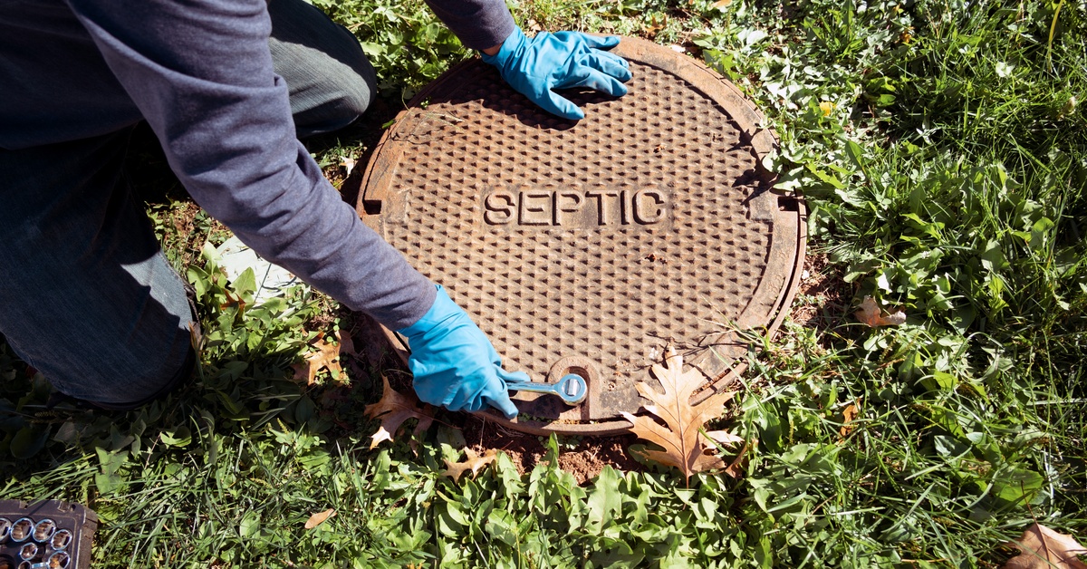 A plumber wearing blue plastic gloves uses a small wrench to unlatch a septic tank's lid from the ground.