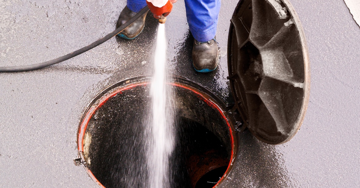 A sewage worker uses a high powered pressure washing hose to blast water into an open manhole in the ground.
