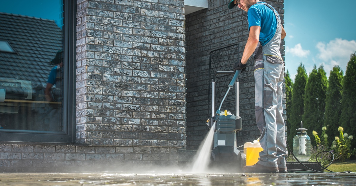 A professional pressure washer in overalls uses a pressure washing machine to blast the ground beneath him.