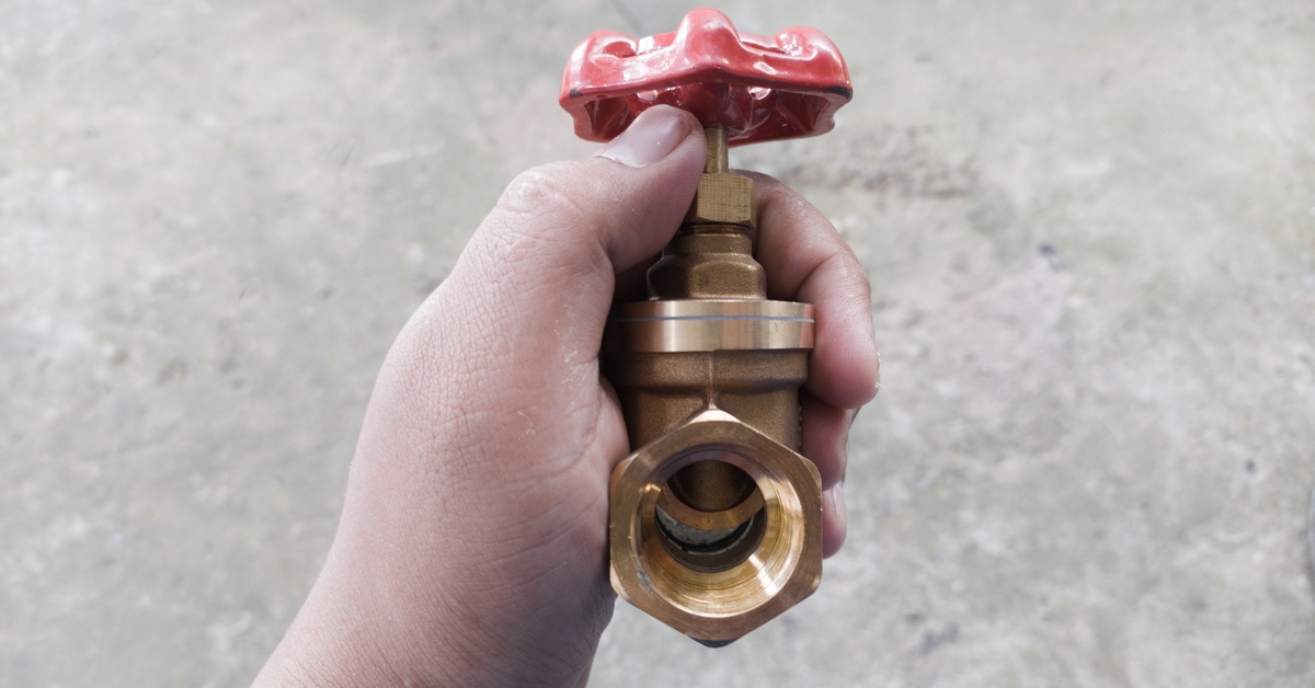 A person's weathered hand holding a brass valve. The brass valve has a red knob on the top of it's casing.