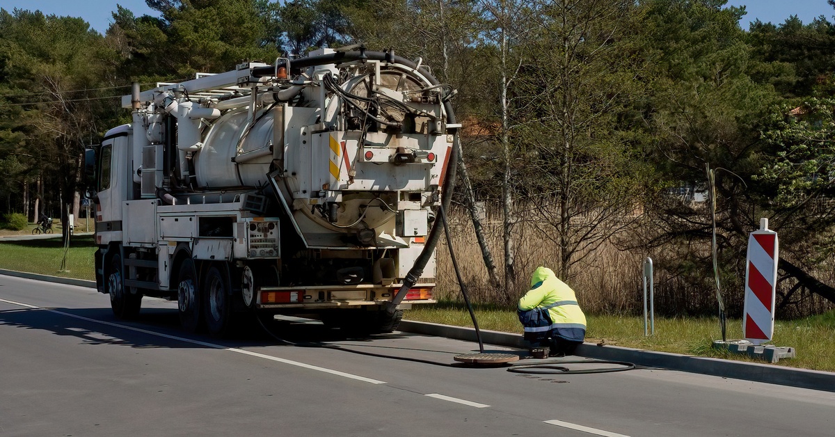 A person in a safety jacket uses a hose attached to a nearby sewage truck to clean out a sewer entrance in the street.