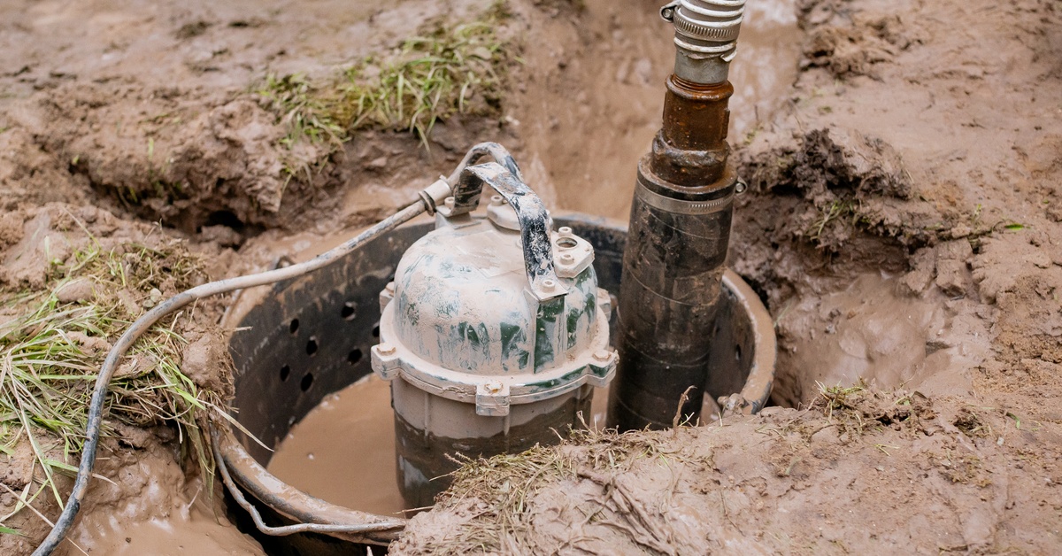 A submersible water pump halfway submerged into a pool of brown water that is coming up from a well in the ground.