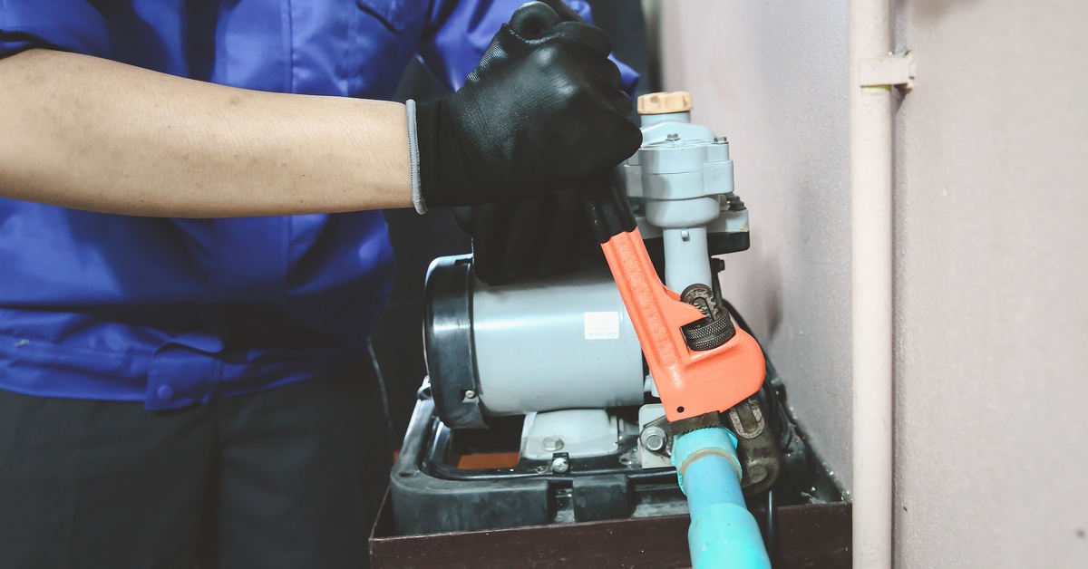 A plumbing technician using a large orange wench to do repairs on a water pump that has a blue pipe coming out of it.