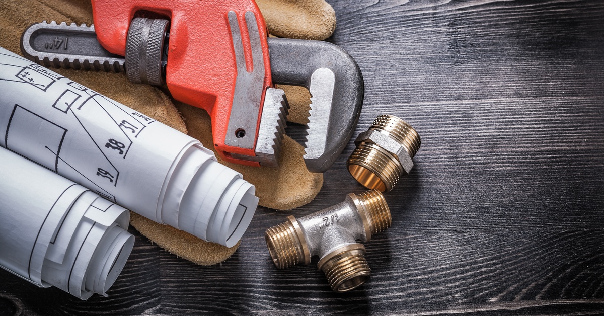 Two small pipe fittings, a pair of gloves, a wrench, and some rolled up blueprints against a wood background.
