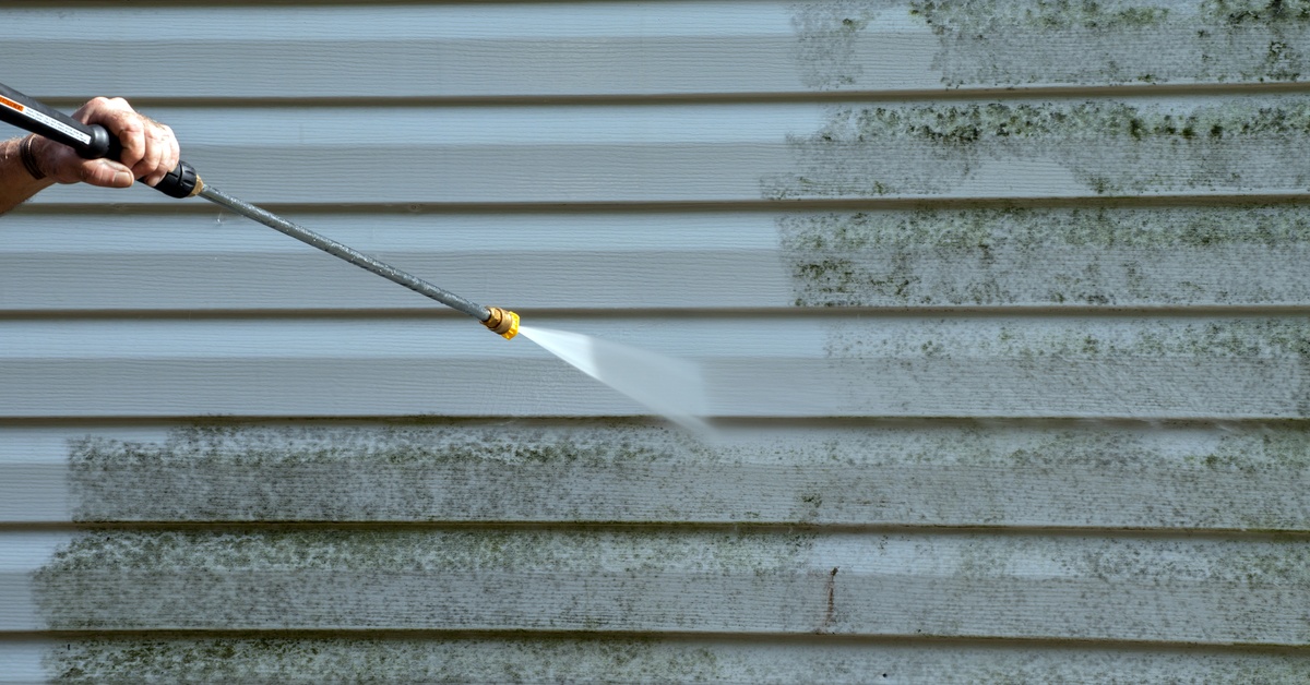 A hand holding a pressure washing hose using the pressurized water stream to blast off dirt from some vinyl siding.