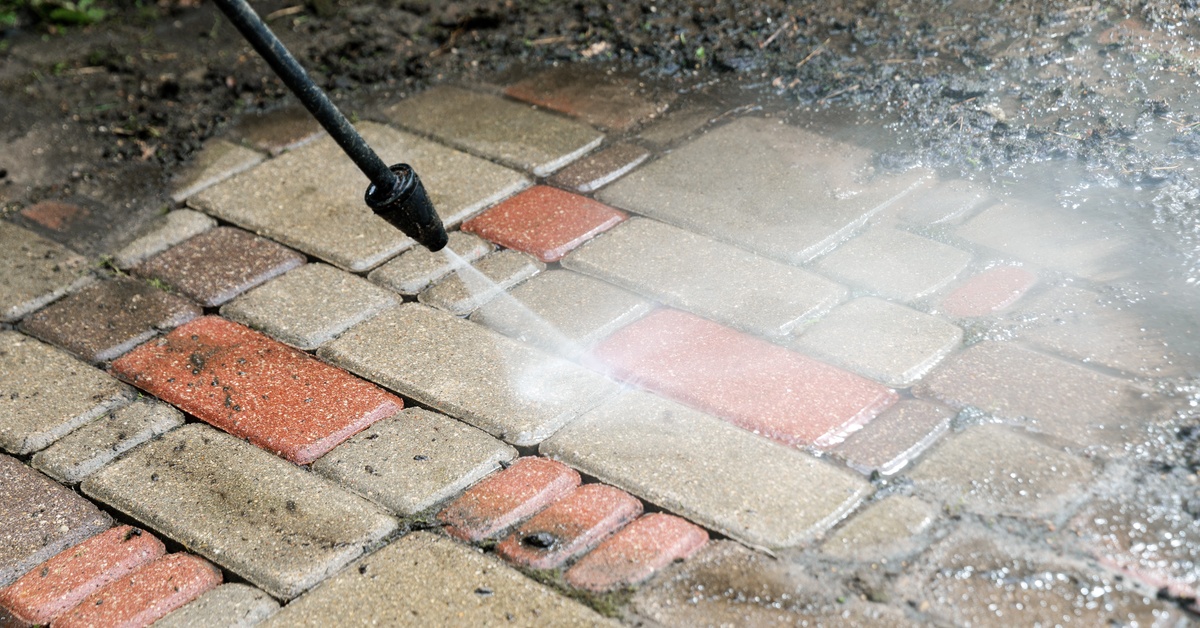 The nozzle of a pressure washing attachment blasting a stream of water to clean off grime from a brick walkway.