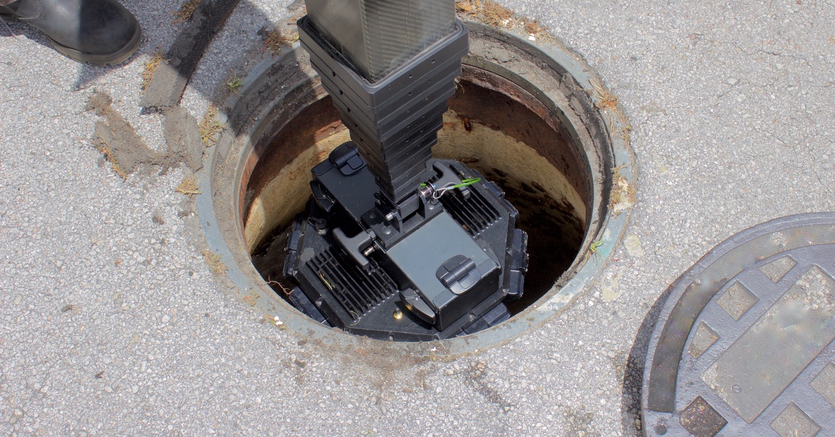 A large digital sewer camera attached to a big, rectangular pipe. The camera is going down an open manhole.