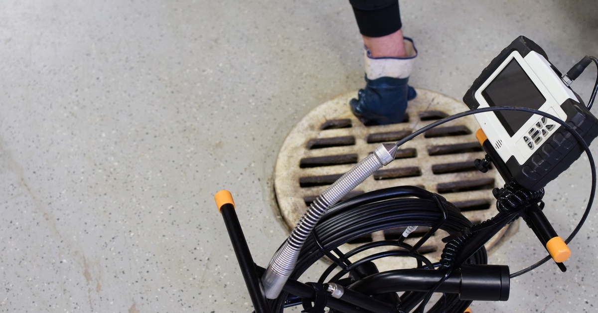 A black and white sewer inspection camera. It is on a stand that is next to a sewer grate in the ground behind it.