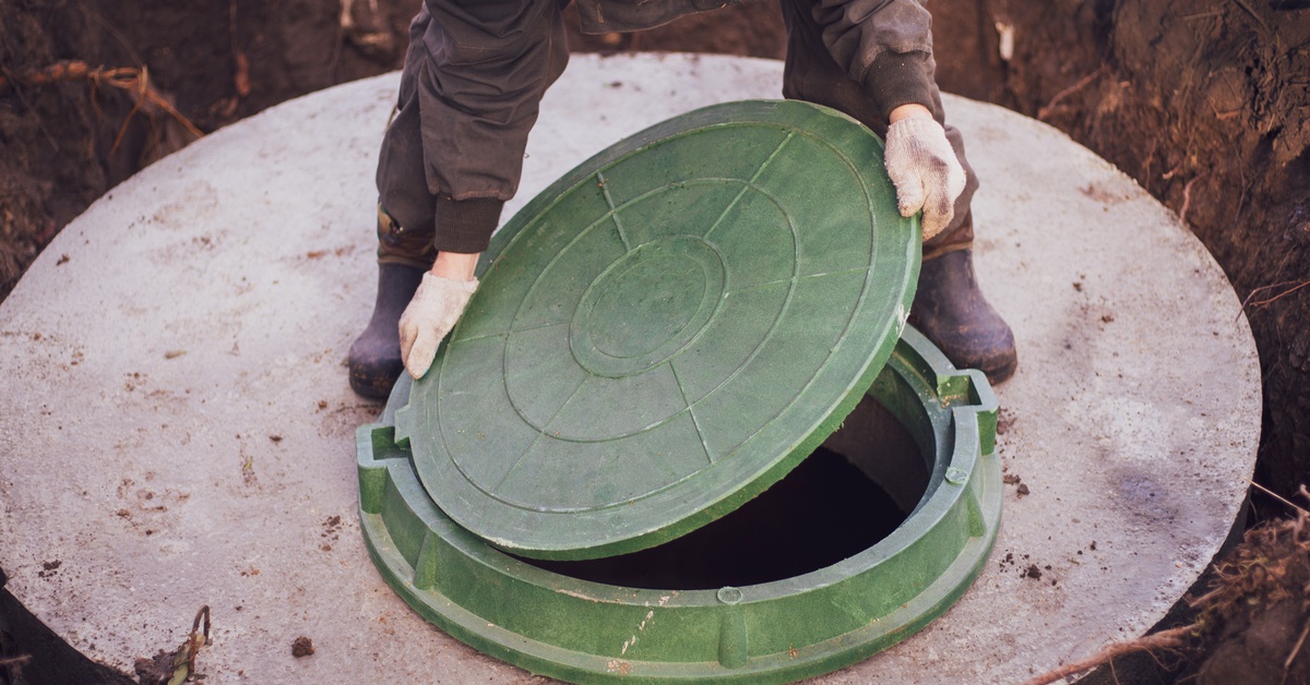 A worker in a coverall lifting up the cover of a manhole. The manhole is built into a concrete slab in the ground.
