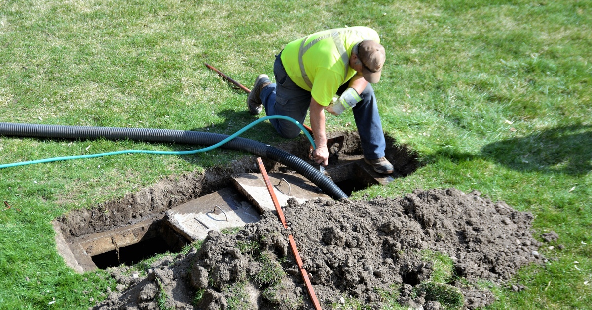 A worker in a yellow safety vest using a large hose to pump out a septic system that is embedded in the ground.