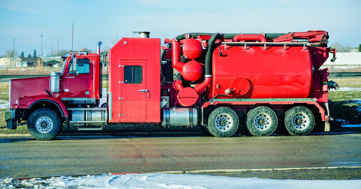 A very large, red hydro excavation truck parked on the side of a road. A stream of soapy water runs in front of it.