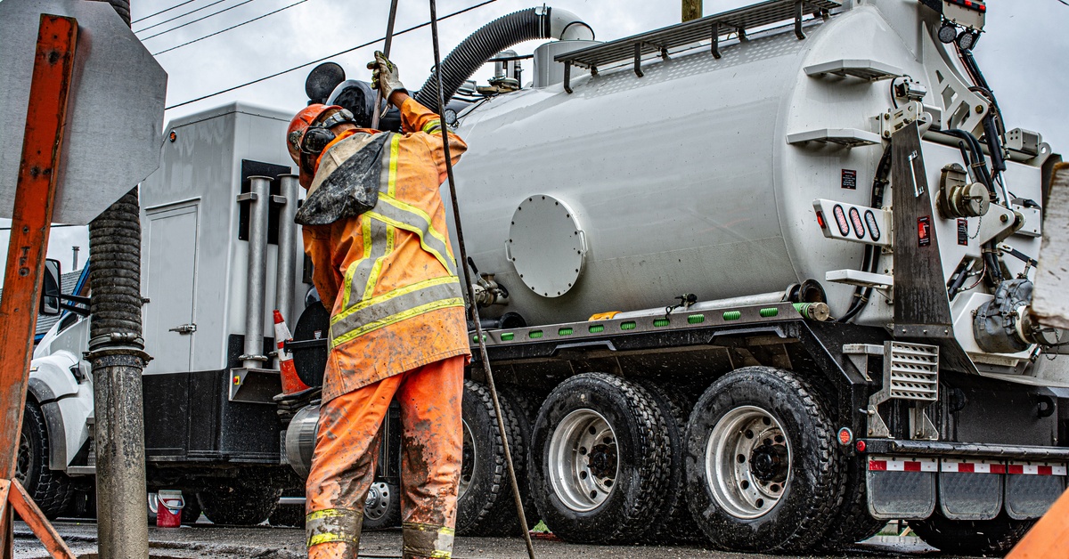 A worker in dirty safety gear using a long pipe to hydro excavate a manhole in the ground. A large truck is behind him.