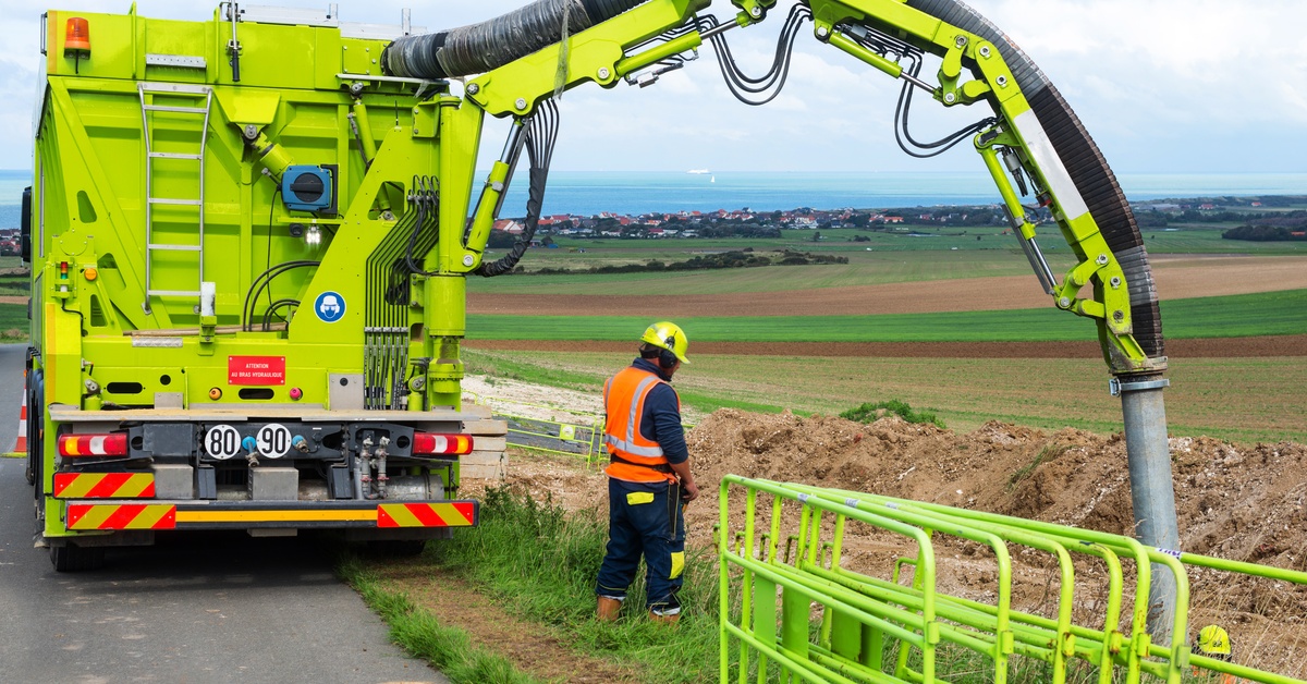 A large, yellow hydro excavation truck with a wide hose coming out of the top of it. A worker stands near the hose.