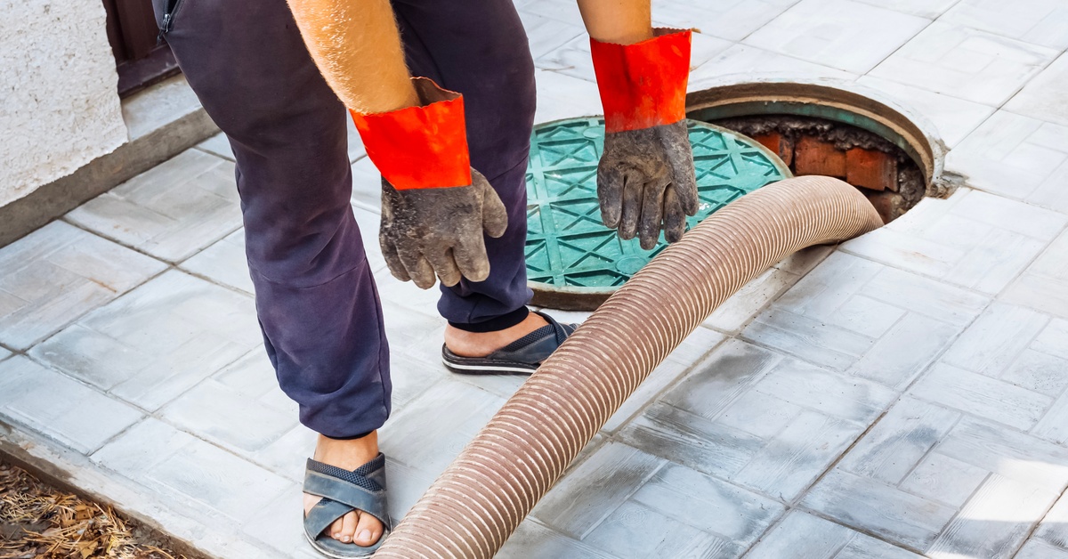 A sewage worker wearing heavy gloves placing a large hose down into an open manhole with its cover next to it.
