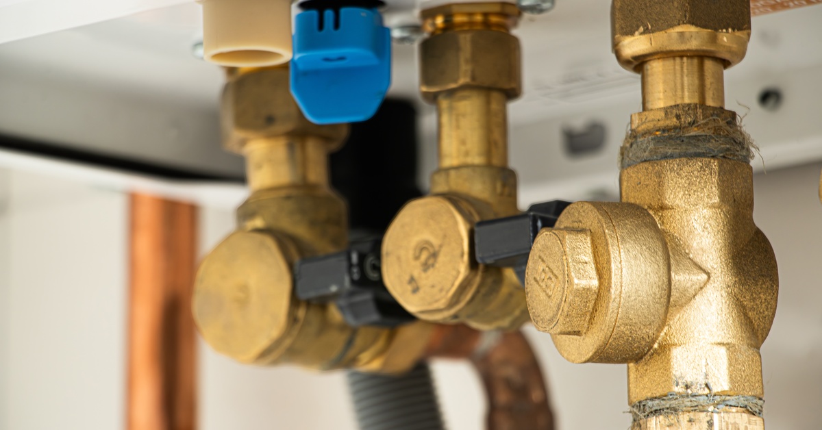 Three old, brass valves that are connected to a plumbing network of pipes underneath a porcelain sink.