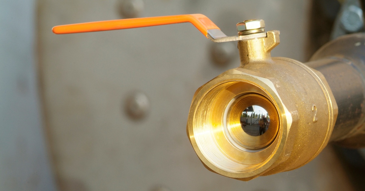 A closeup on a brass ball valve that is part of a large pipe system. The valve has an orange handle on it.