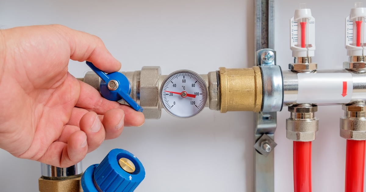 A man's hand turning a blue, plastic valve on the side of some pipes. There is a small gauge in the pipe's center.