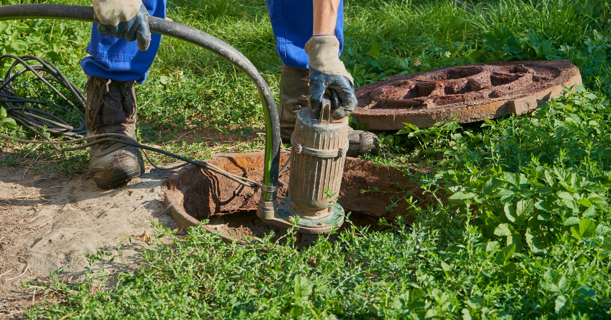 A plumber in blue jeans using a vacuum pump to pump water out of cesspool that is behind someone's house.