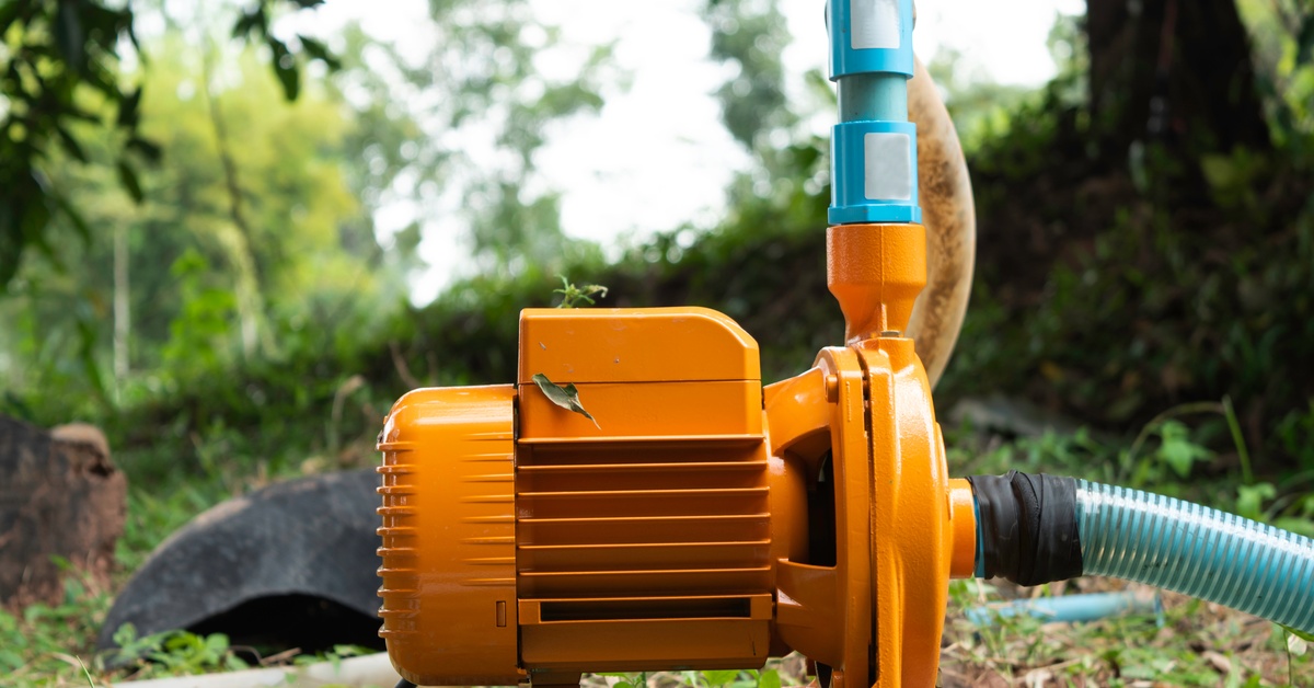 An orange water pump set up on a rock in the middle of some grass. Two separate hoses are coming out of the pump.
