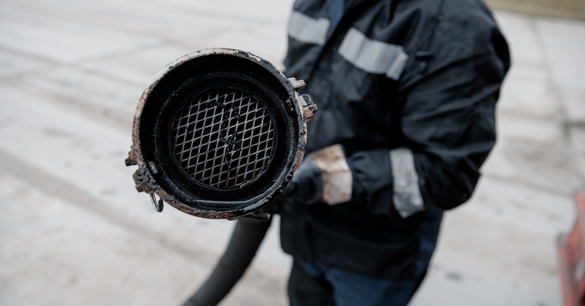 A sewage worker holding a large pipe and connector. He is pointing the hose's end at the viewer of the picture.