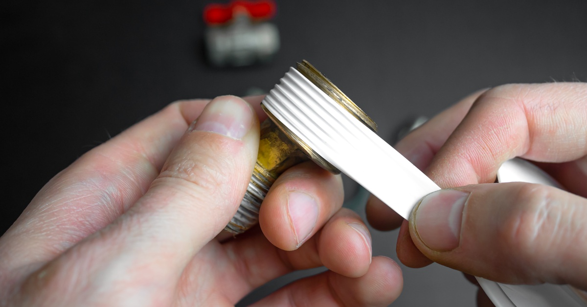 The hands of a plumber stretching a small section of plumber's tape over the threaded end of a pipe connector.