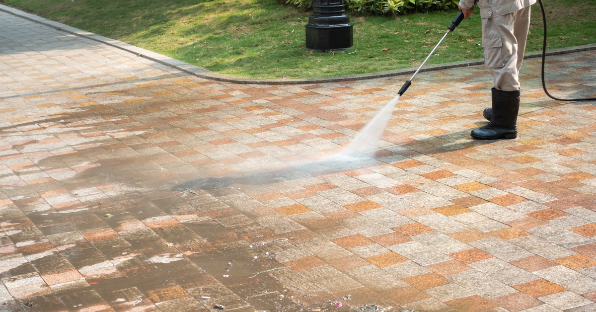 A person in a jumpsuit and rubber boots using a pressure washer to blast dirt off of the brick path on the ground.