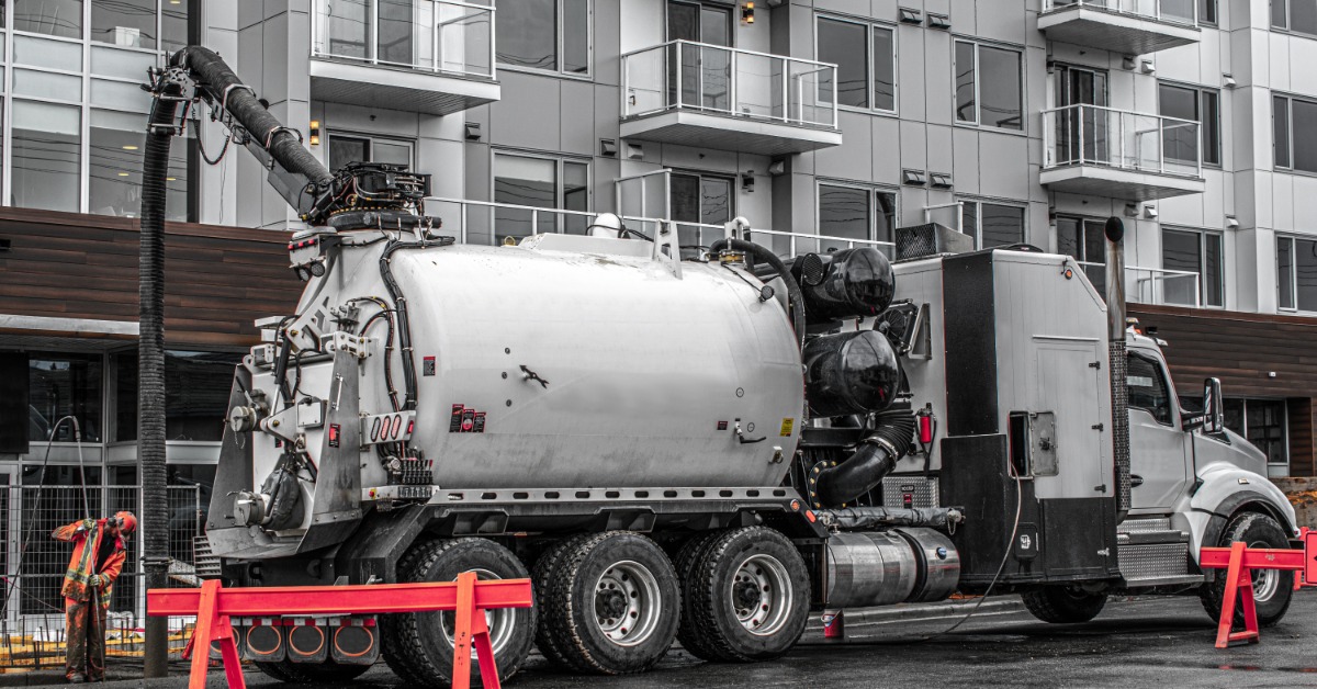 A large hydrovac truck parked outside an apartment building. A worker in orange is using a hose connected to it.