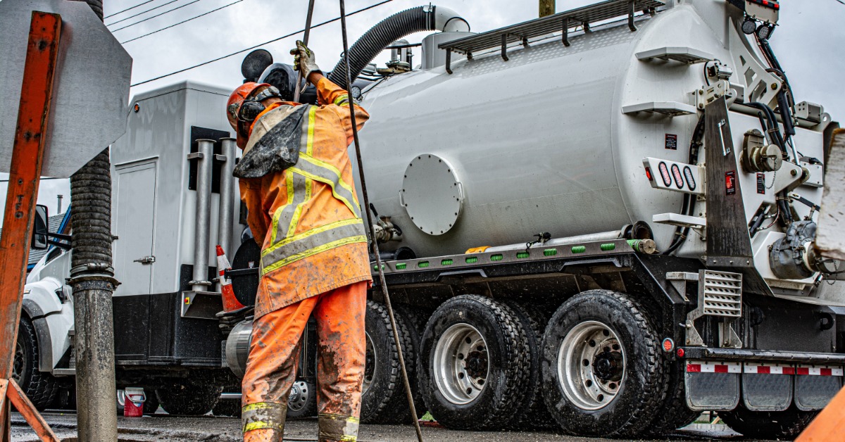 A hydrovac worker in orange high-visibility gear using a large hose connected to the truck parked behind him.