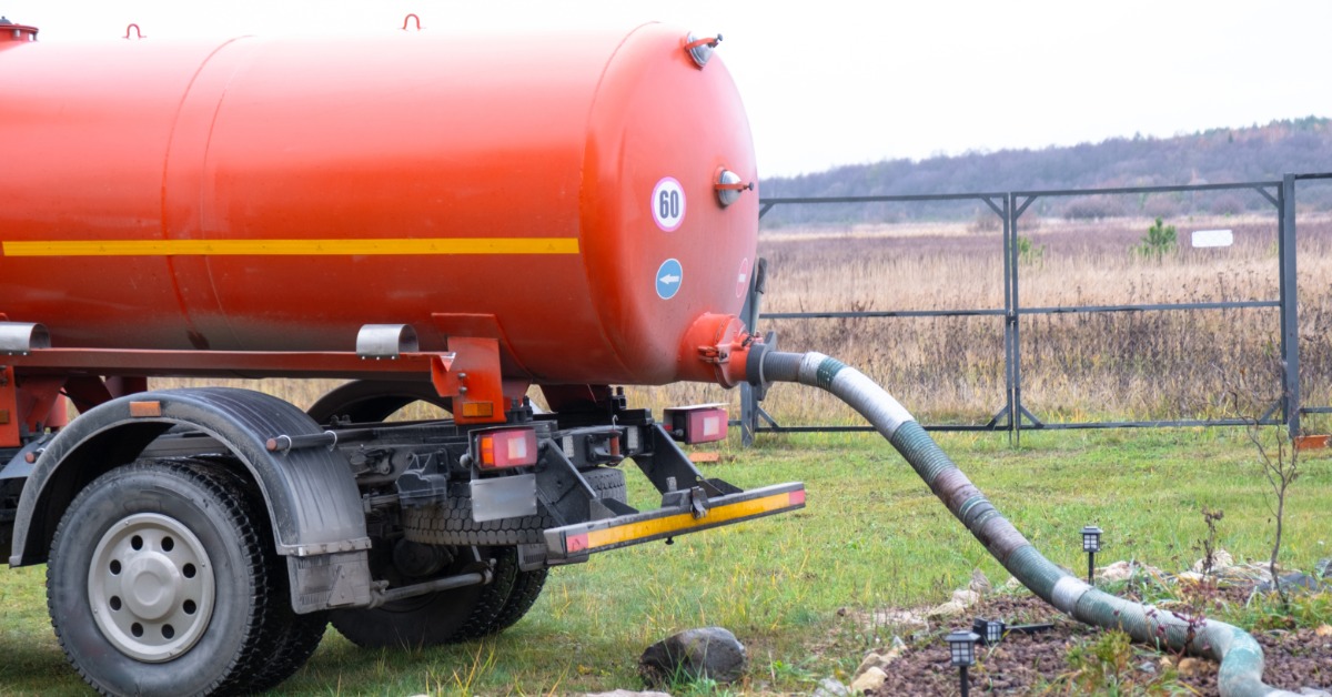 A large, orange sewage truck parked on the grass. A wide hose is coming out of the back of the tank on the truck.