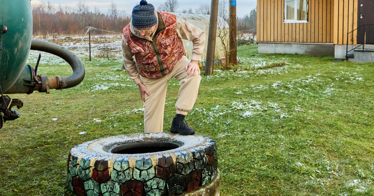 A sewage worker wearing a camouflage vest looking down into a cesspit while standing next to a vacuum truck.
