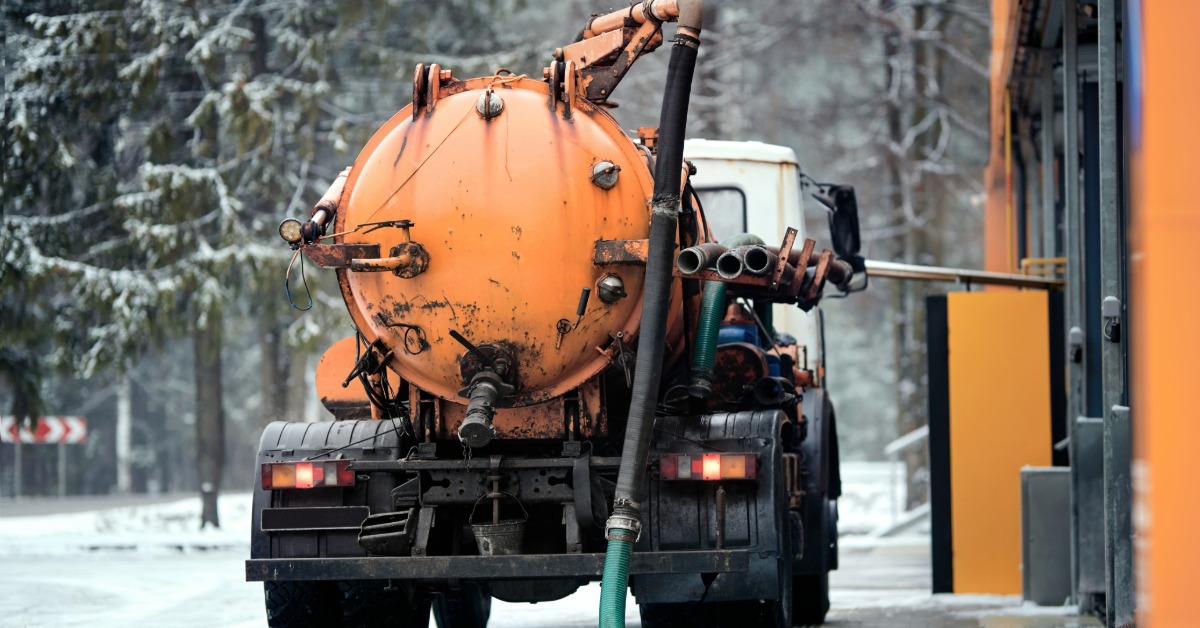 An orange vacuum pump truck parked outside in the snow. A hose travels from the top of the truck and off to the side.