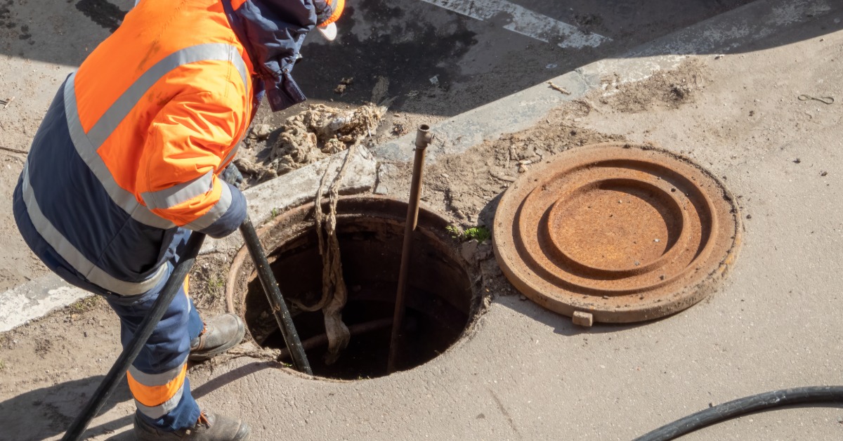 A sewer worker wearing an orange safety jacket feeding a hose down into an open manhole in the street.