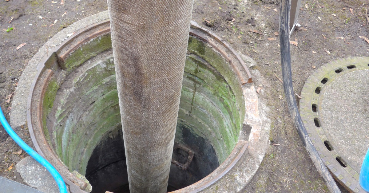 A manhole with its cover off and off to the side. A large, wide hose is being fed into the open manhole.