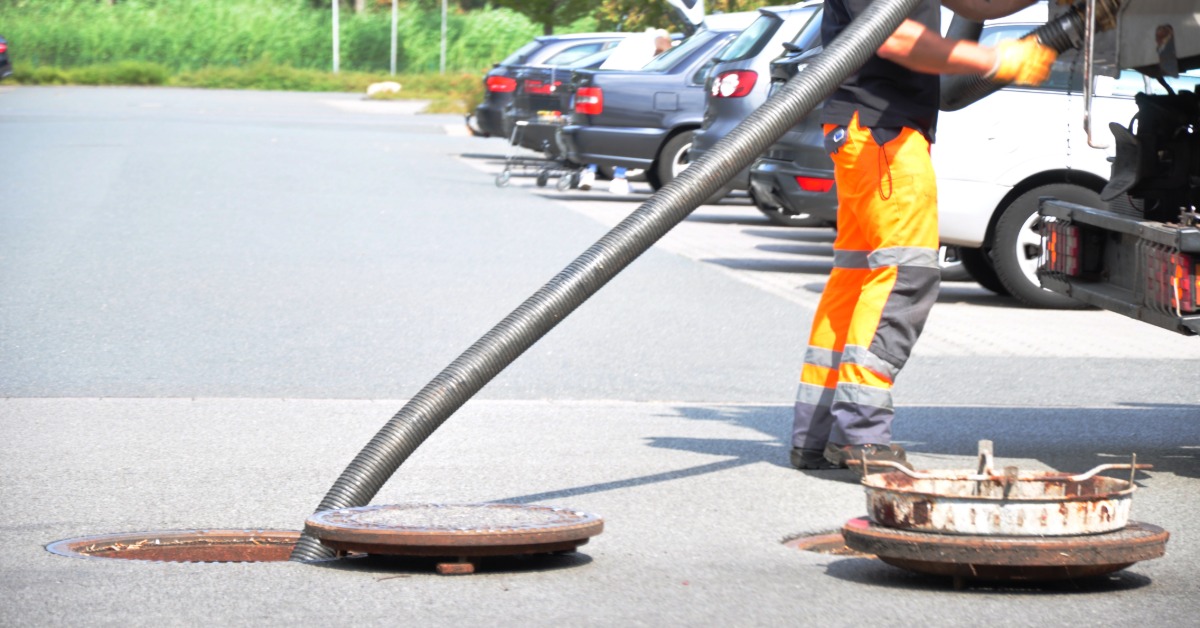 A sewer worker in orange safety trousers standing next to an open manhole with a large hose going into it.
