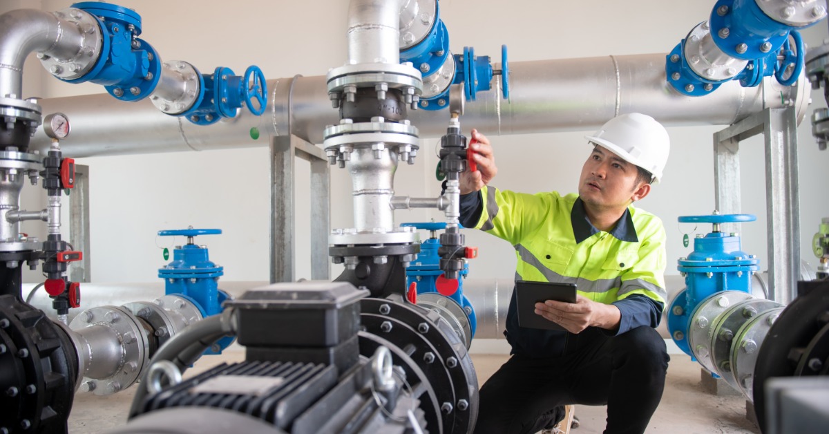 A mechanic in a yellow safety vest inspecting a series of pipes and water pumps in a large, white room.