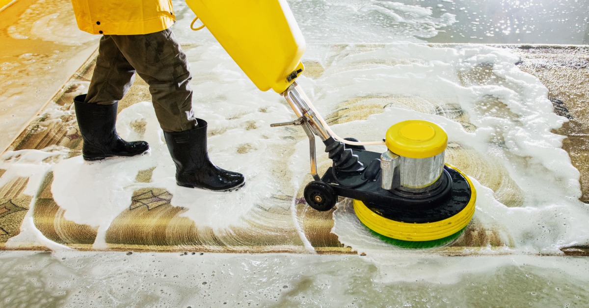 A carpet cleaner in a yellow jacket using a large cleaning machine to scrub away the dirt from a rug below.