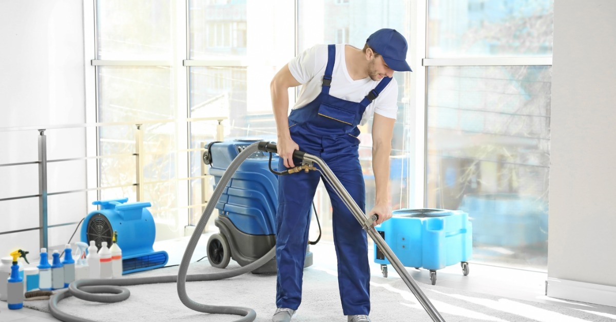 A dry cleaner using a large vacuum cleaner on a rug inside of a home with large windows open just behind him.