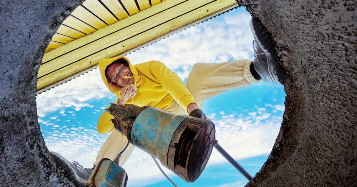 A view from inside a sewer pipe of a man in a yellow sweatshirt about to lower a waste pump into the pipe.